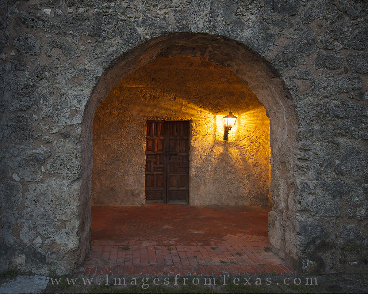 A stone hallway in San Antonio's Mission Concepcion showcases a Colonial style door as it is lit by a lamp.