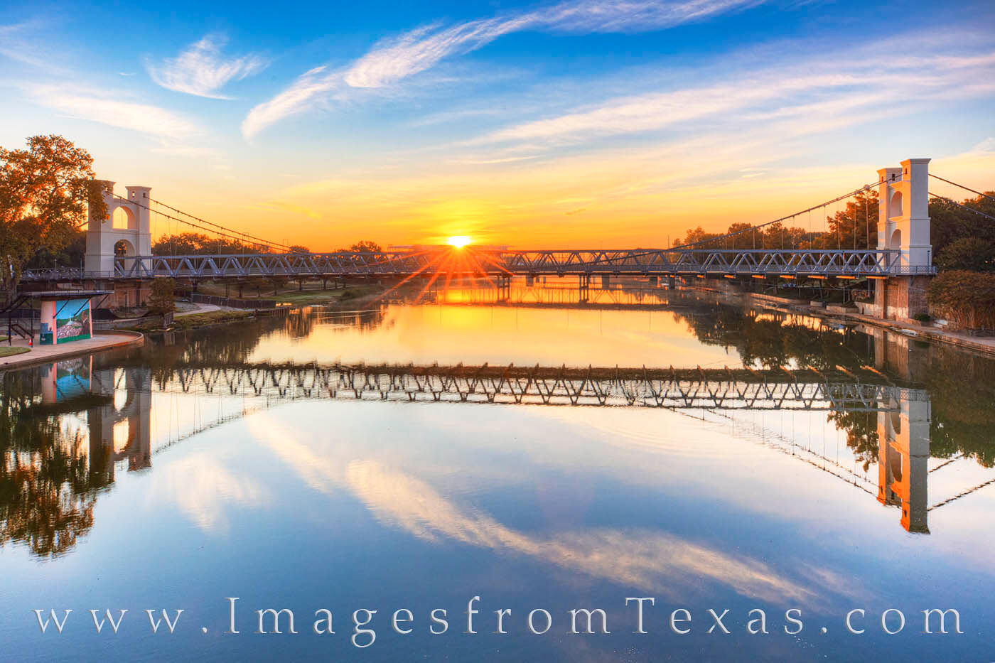 From the Washington Avenue Bridge on the Brazos River, this view shows the famous Waco Suspension Bridge at sunrise. In the distance...